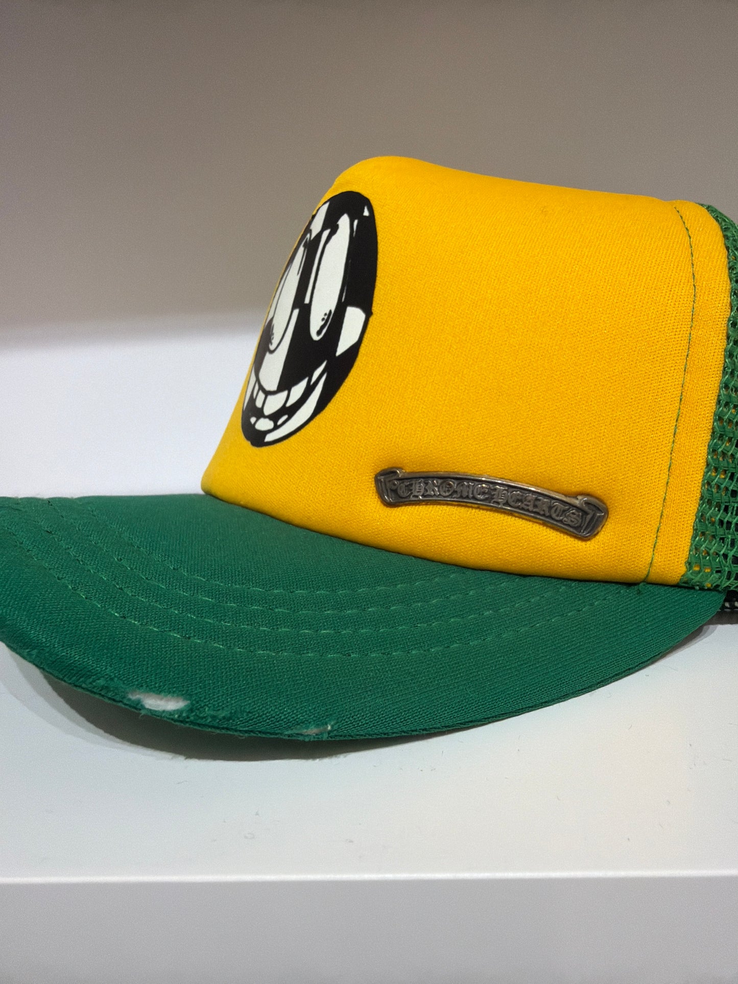 Side angle of Chrome Hearts Matty Boy yellow green trucker hat showing curved brim and silver Chrome Hearts plaque