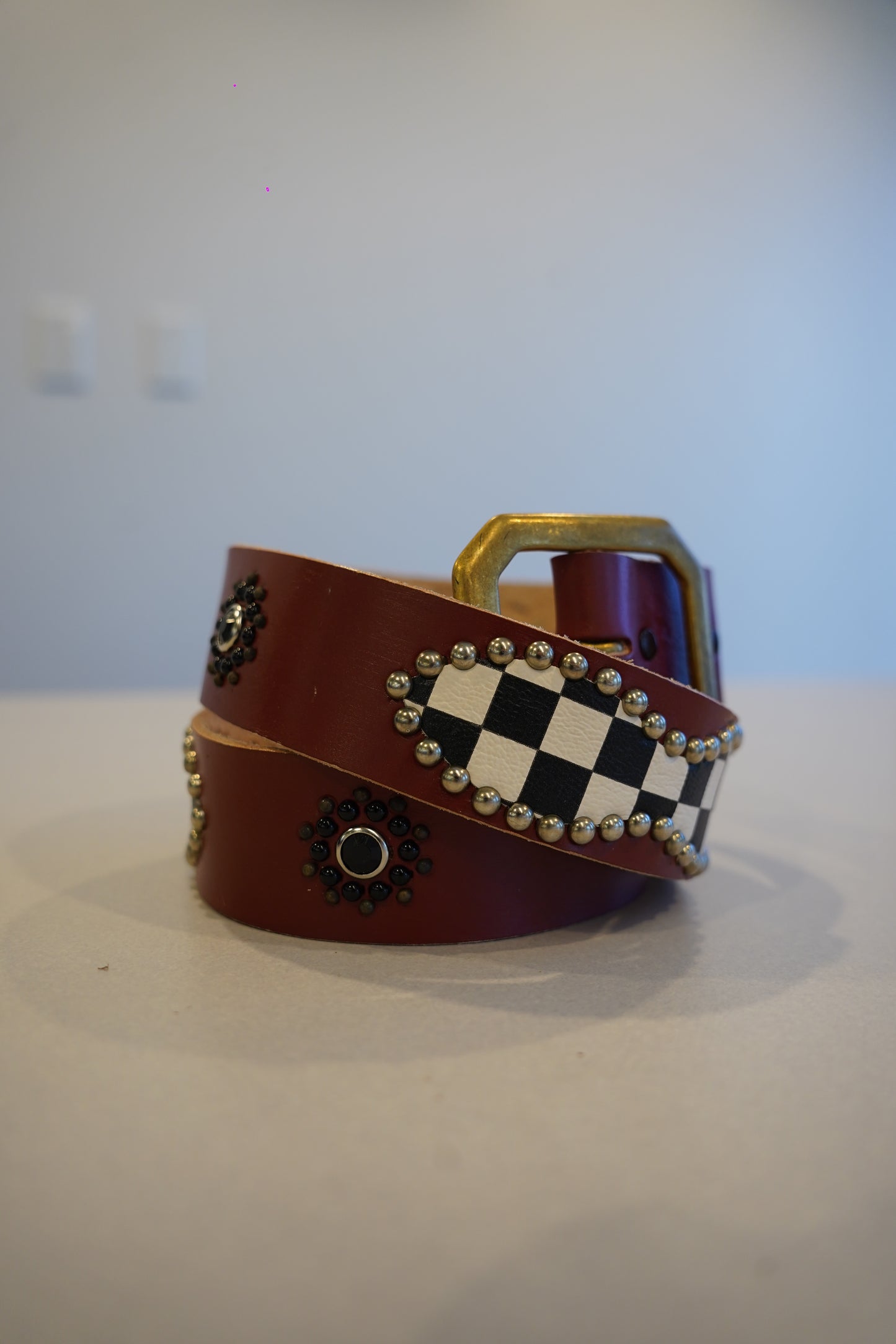 Vintage burgundy leather belt coiled with brass buckle, checkerboard studded panel and beaded rosette accents.