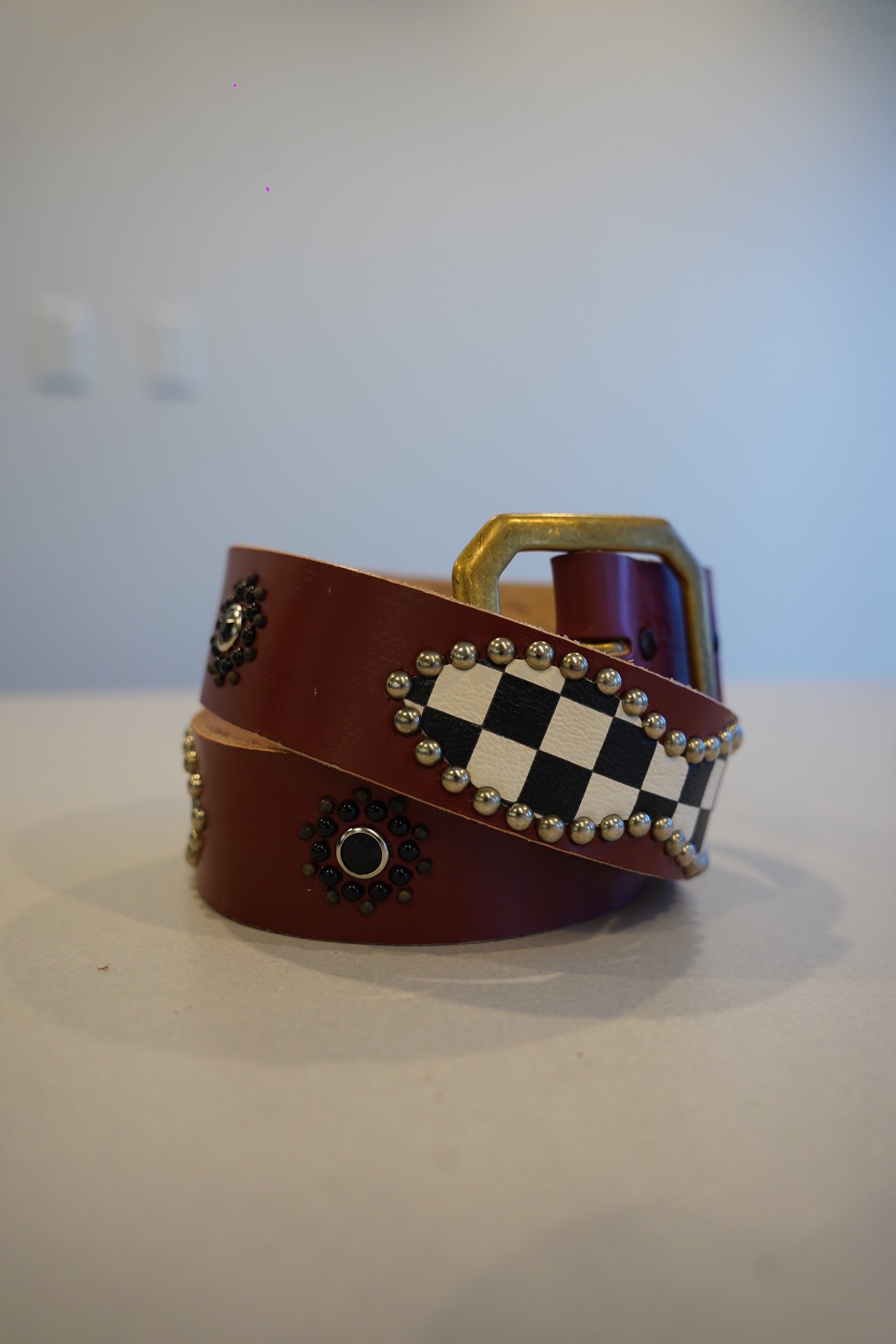 Vintage burgundy leather belt coiled with brass buckle, checkerboard studded panel and beaded rosette accents.