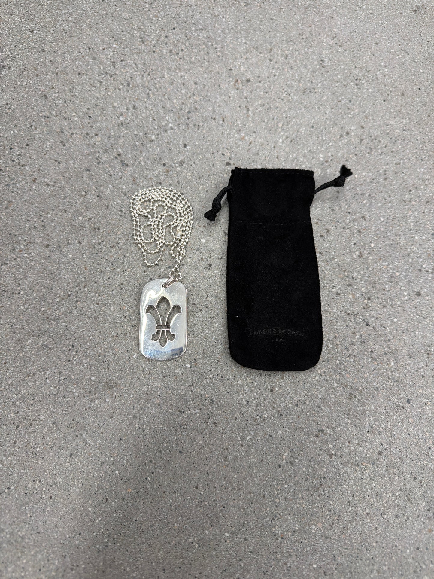 Chrome Hearts Fleur dog tag pendant shown with branded black drawstring jewelry pouch