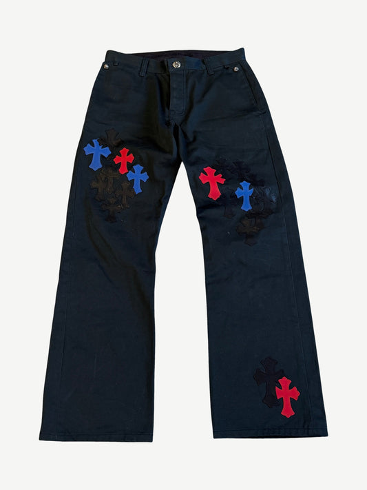Chrome Hearts black chino pants front covered in stitched black, red and blue leather cross patches at thighs.