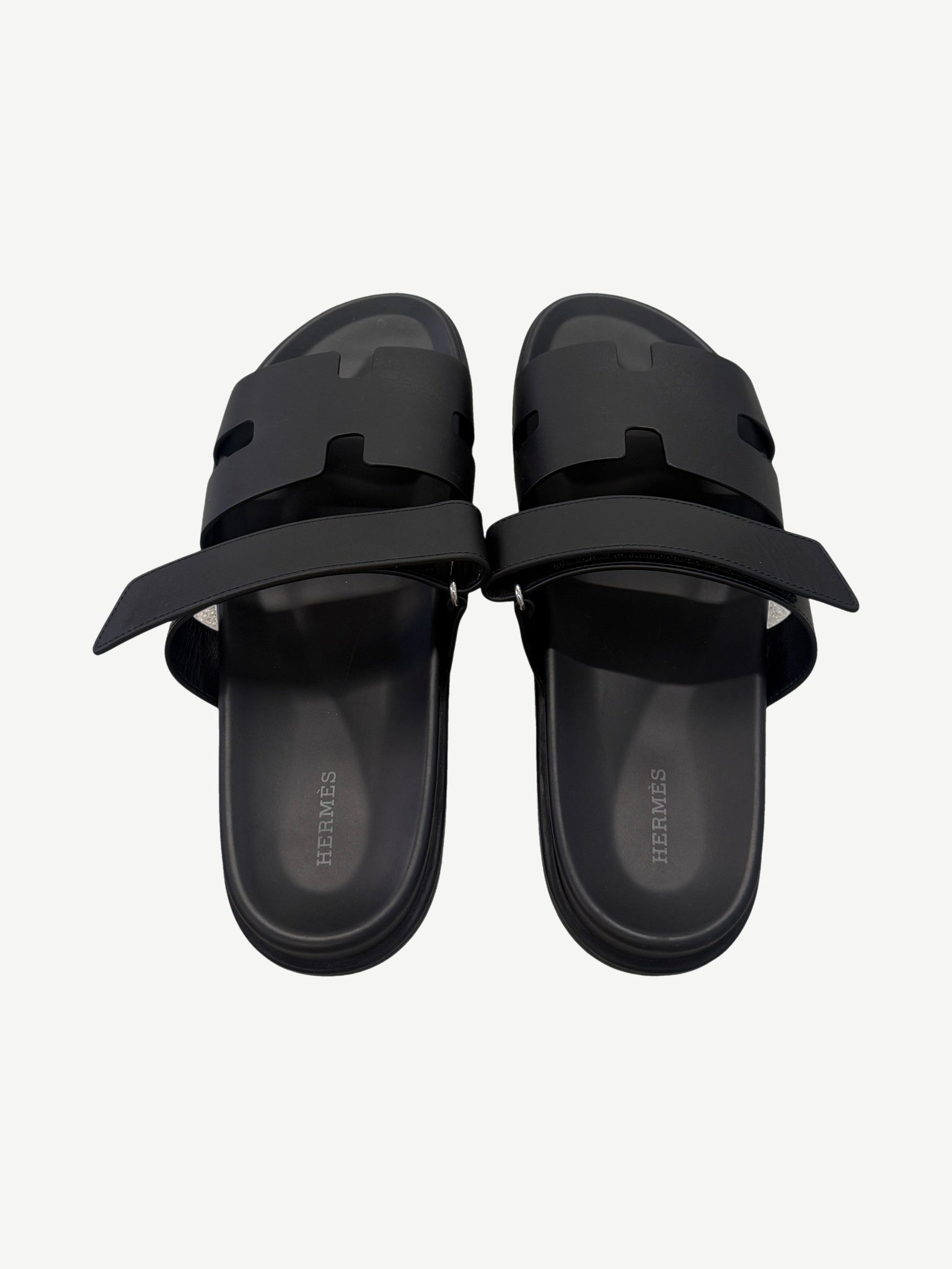 Hermes Chypre sandal pair top-down view, black calfskin straps unfastened revealing Hermes branded leather insoles.