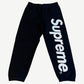 Supreme black satin applique sweatpants front view with iridescent Supreme wordmark down right leg panel