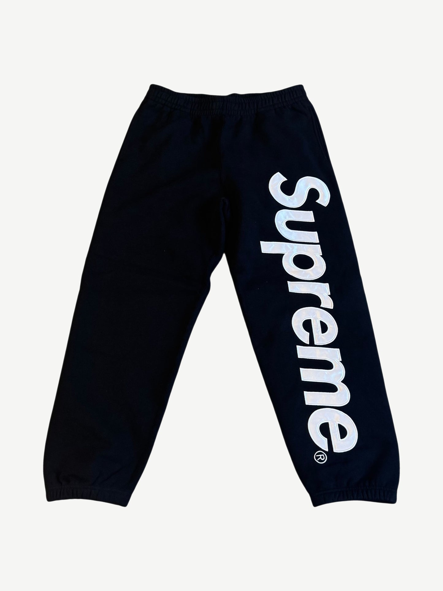 Supreme black satin applique sweatpants front view with iridescent Supreme wordmark down right leg panel